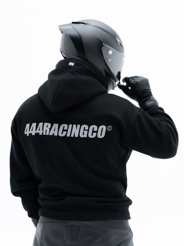 A person from behind wearing a black hoodie with '444RACINGCO' printed in white across the back, a black motorcycle helmet, and black gloves, set against a plain white background.