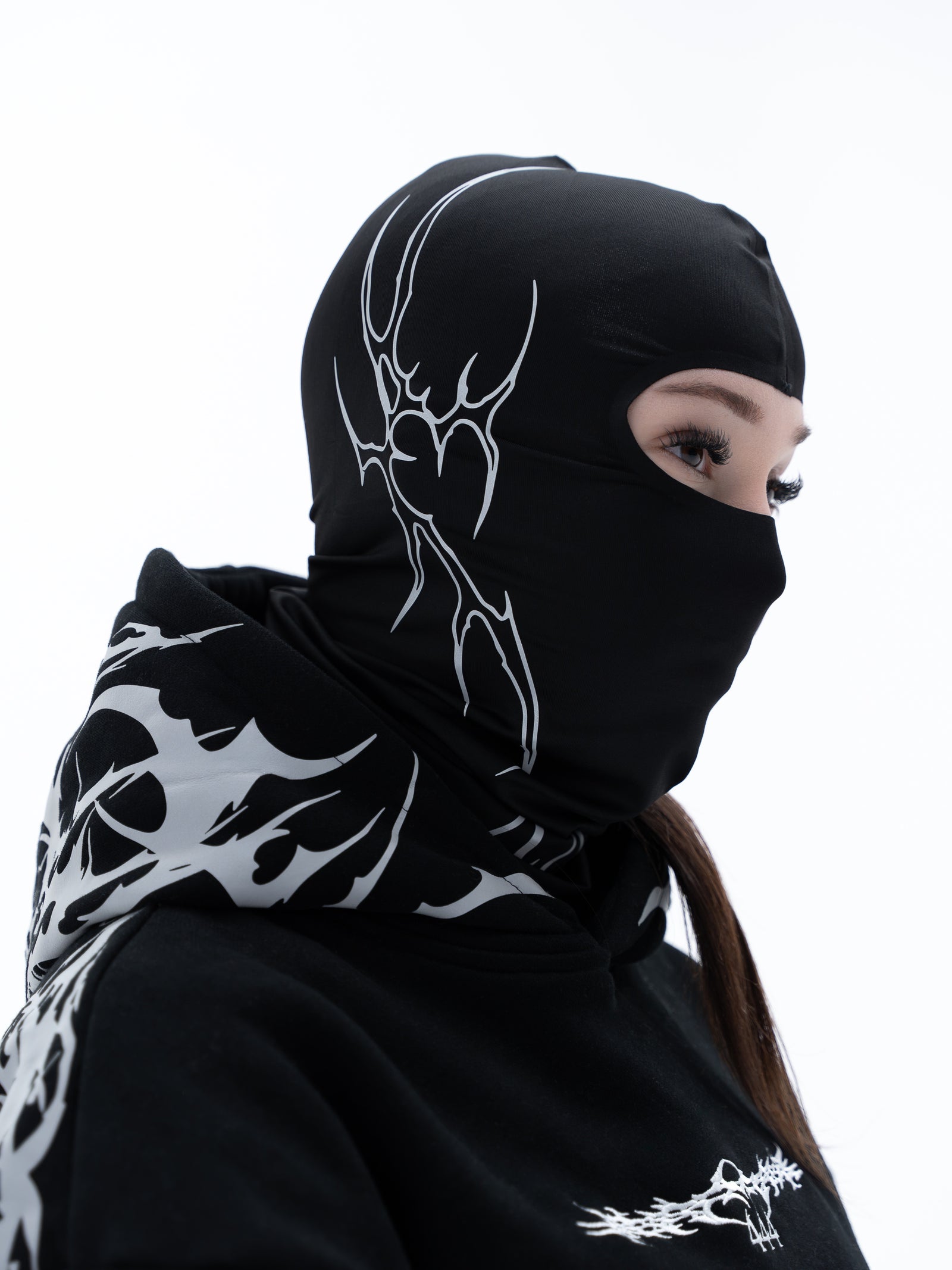 A person wearing a black full-head balaclava mask with white graphic line art and a matching black hoodie with white flame-like graphics, shown against a plain white background.