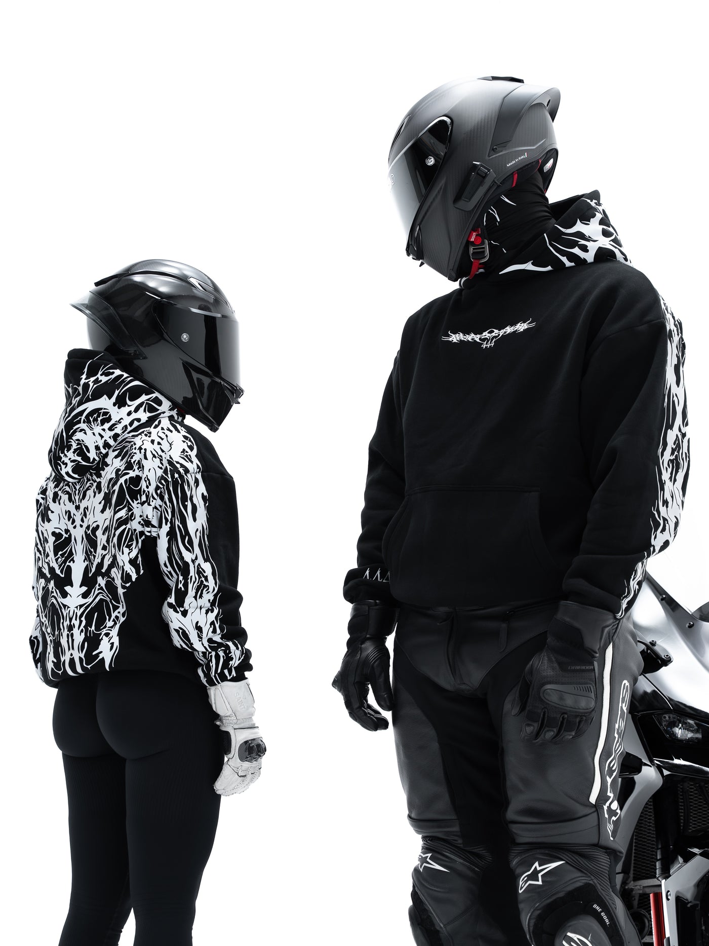 Two models wearing black Alpinestars motorcycle apparel with white flame-patterned sleeves and hood, complete with helmets and riding pants, set against a solid white background.
